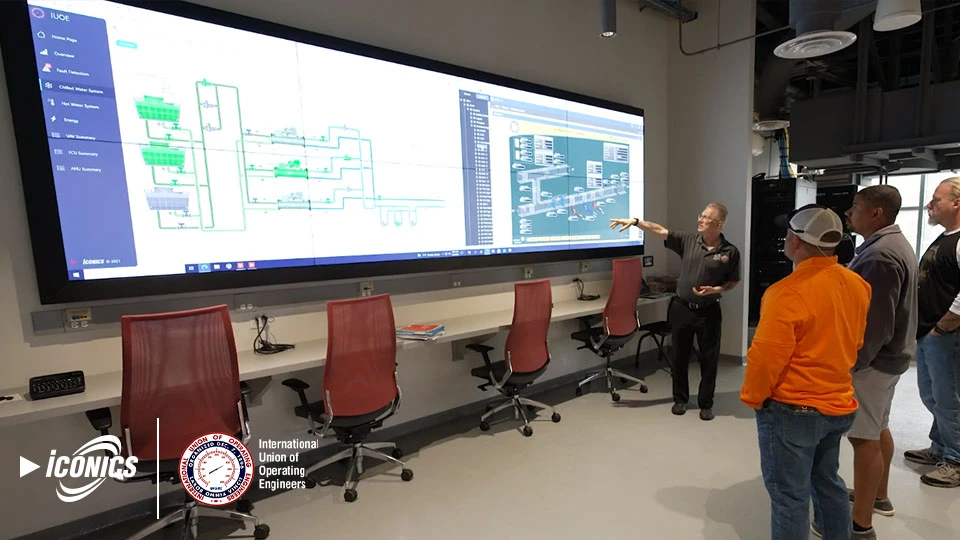 Photo of four men in front of a full-wall length digital display showing pipeline, flow and valve schematics